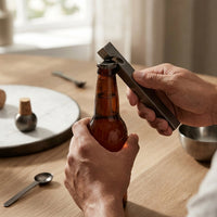 Person opening a beer bottle with a bottle opener on a wooden table.