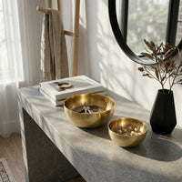 Decorative setup with gold bowls, a black vase with dried plants, and books on a stone surface.