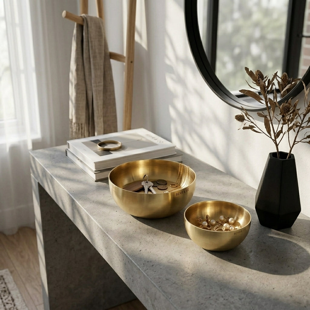 Decorative setup with gold bowls, a black vase with dried plants, and books on a stone surface.