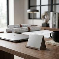 Modern desk setup with laptop, mug, business card holder and books in a bright room.