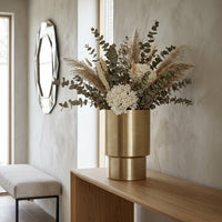 Gold vase with flowers on a wooden console table in a room with a mirror and bench.