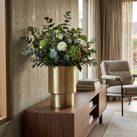Decorative bouquet in a gold vase on a wooden console table with a chair and window in the background.