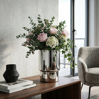  Bouquet of flowers in a silver planter on a wooden table with a chair and books in the background.