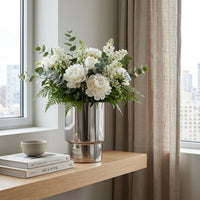 Floral arrangement in a silver vase on a wooden surface with books and a cup.