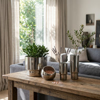 Metal vases, metal pot, metal salt holder on a wooden table in a living room with plants and vases.