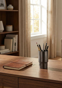 Home office desk with a notebook, pen holder, mug, and plant in a well-lit room.