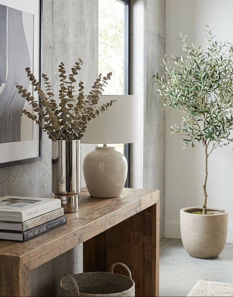 Decorative setup with a wooden console table, books, a vase, and a potted plant in a room with large windows.