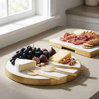 Round marble cheeseboard with gold rim on a light wooden floor, featuring cheese, fruits, and meats.