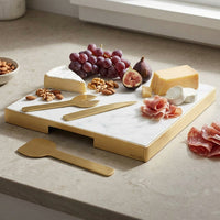 Marble cheeseboard with gold cutlery, cheese, fruits, and meats on a light stone surface.