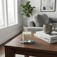 Candle placed on a metal coaster on a wooden table with a cup and books in a cozy living room.