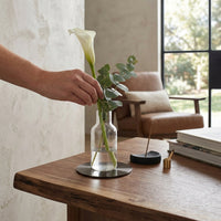 Person arranging flowers in a clear vase on a black metal coaster on a wooden table with a blurred background