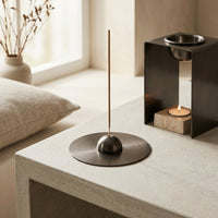 Incense holder holding an incense stick on a metal coaster with an oil burner in background