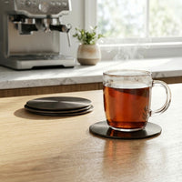 Clear glass mug with steaming hot liquid on a black nickel coaster in a kitchen setting with a coffee machine and window.