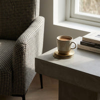Cup of coffee on a metal coasters next to a patterned chair with a window in the background.