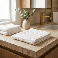 Folded cotton facial towel on a bathroom counter with a vase of greenery and gold faucet.