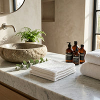 Bathroom counter with stone sink, white facial towels, and bottles on a marble surface.