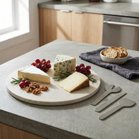 Cheese platter on a stone cheese tray with assorted cheeses, grapes, and nuts on a kitchen counter with metal cheese knives.