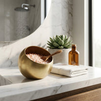 Gold bowl with pink Himalayan salt, wooden spoon, and bottle on a marble surface in a bathroom.