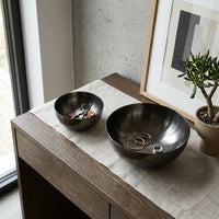 Two black bowls on a wooden surface with coins and rings, next to a plant and framed art.