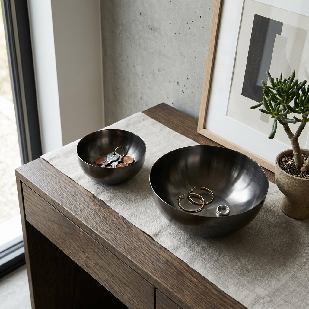 Two black bowls on a wooden surface with coins and rings, next to a plant and framed art.