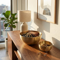 Wooden console table with brass bowls, lamp, and plant in a bright room.